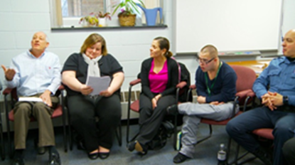 a group of individuals sitting in chairs discussing and listening. One woman is looking at a paper.